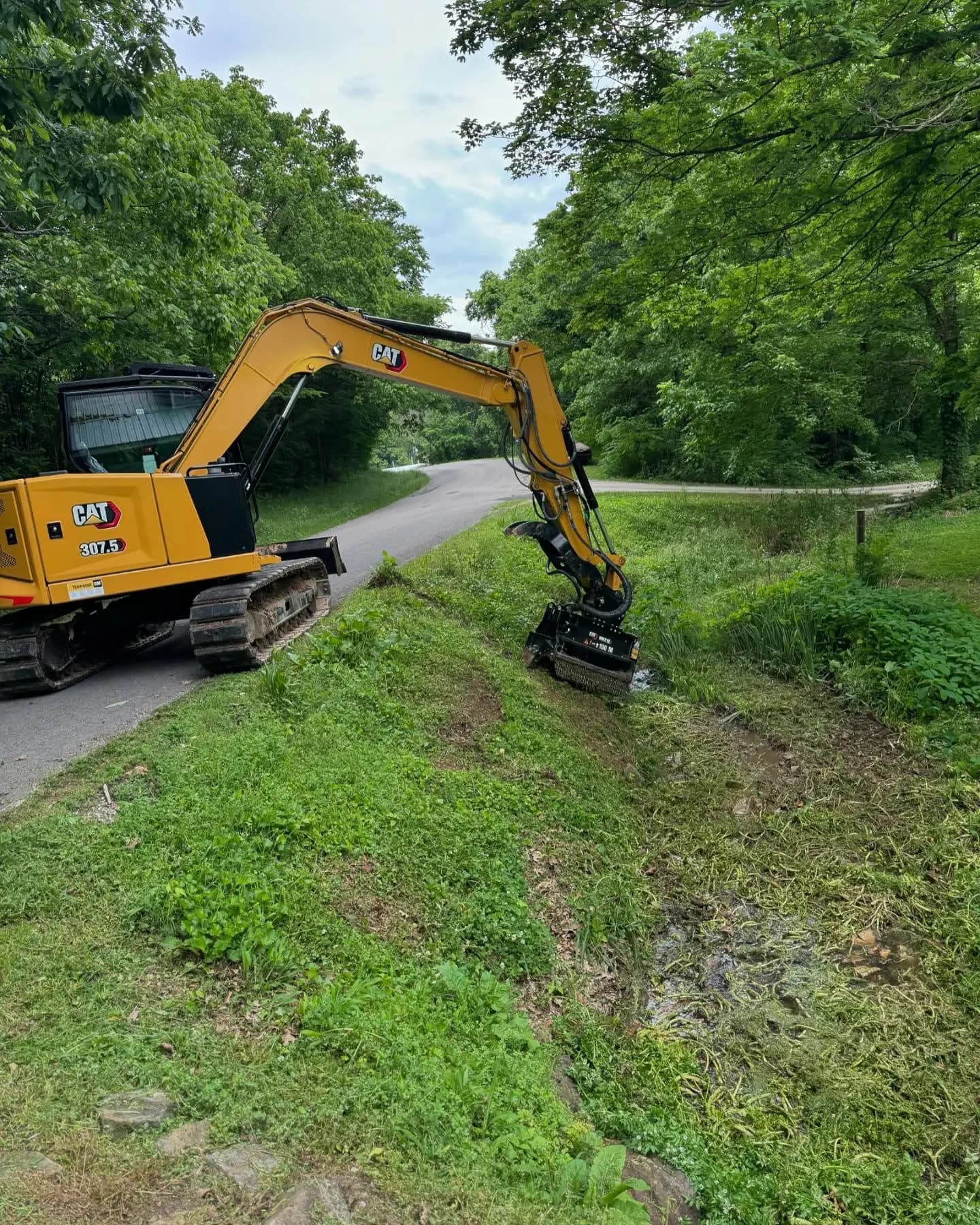 Excavator mulching a roadside ditch bank beside a paved lane