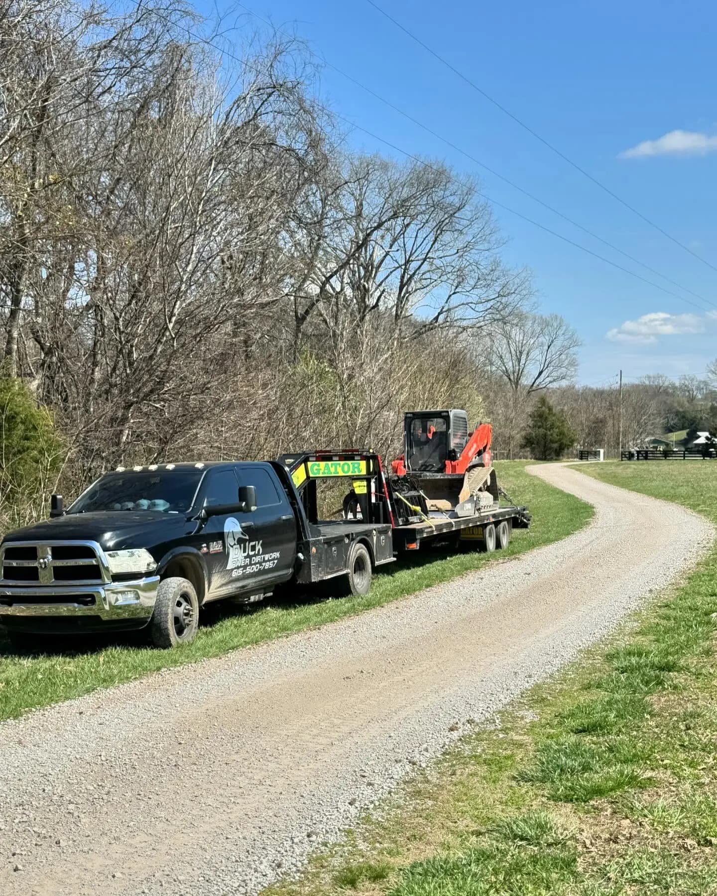 Duck River Dirtwork truck hauling a track loader on a gooseneck trailer along a gravel drive