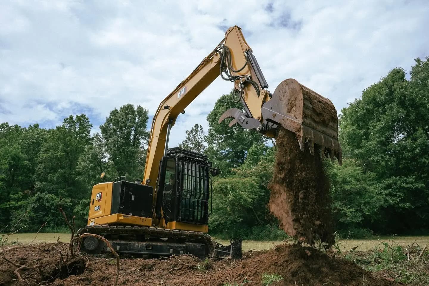 CAT excavator with a grapple dropping a bucket of soil on a cleared field